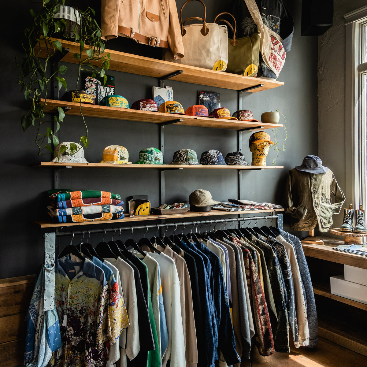Interior photo of a Kapital display inside Canoe Club. There are wooden shelves lining the wall with Kapital Trucker hats and a green ivy plant. The lower rack ha hanging items from Kapital like button up shirts and jackets.