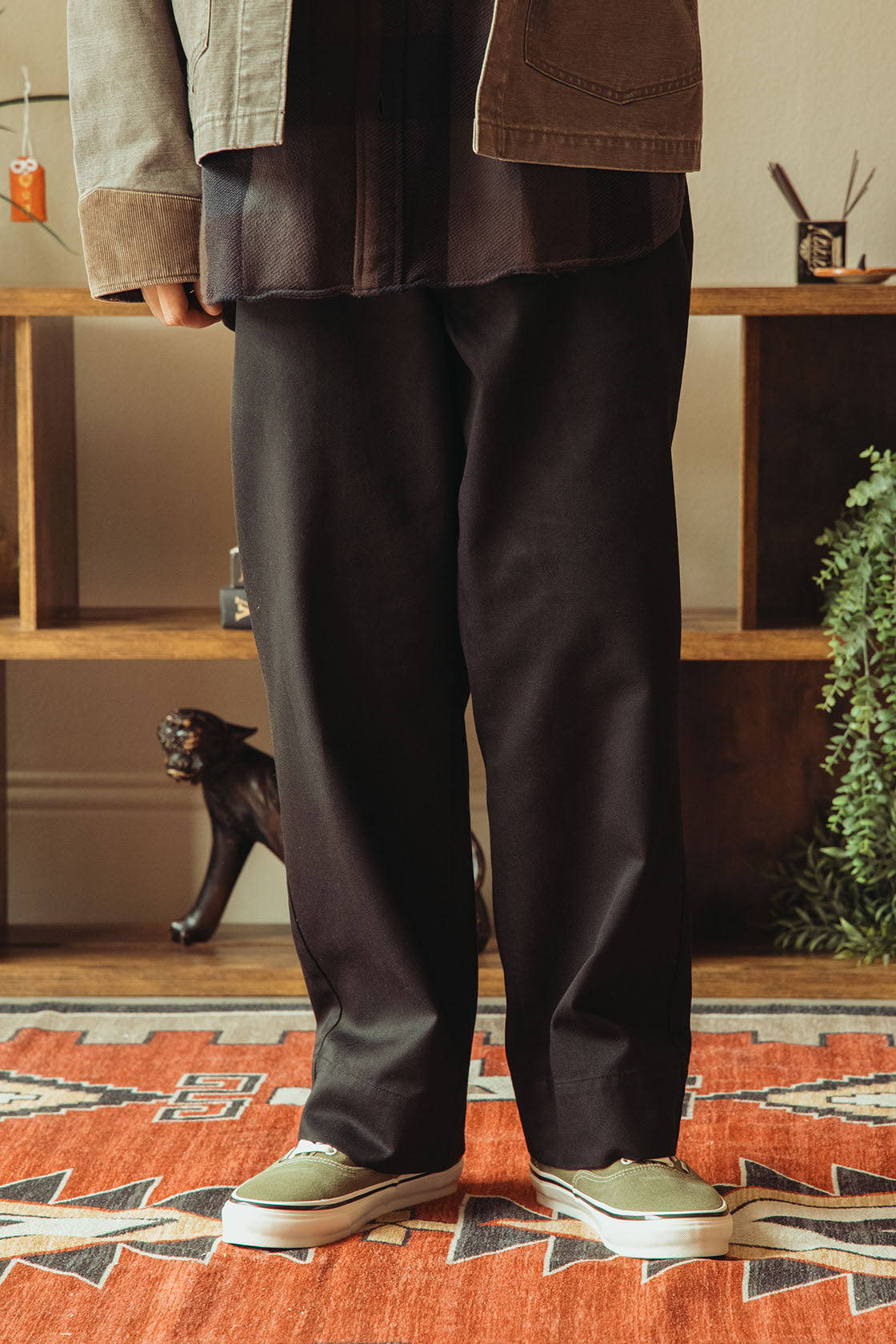A person shown from the waist down wearing loose black wide-leg trousers, a washed black jacket, and olive green sneakers, standing on a patterned rug in front of a wooden shelf.