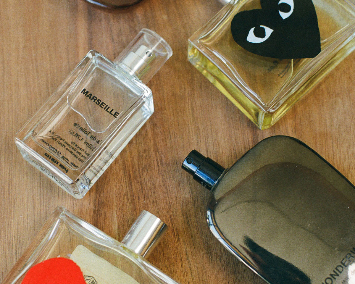 Several perfume bottles arranged on a wooden surface, including a clear rectangular bottle labeled MARSEILLE, a square bottle with a black heart-shaped label, a dark reflective bottle, and partial other bottles at the edges.