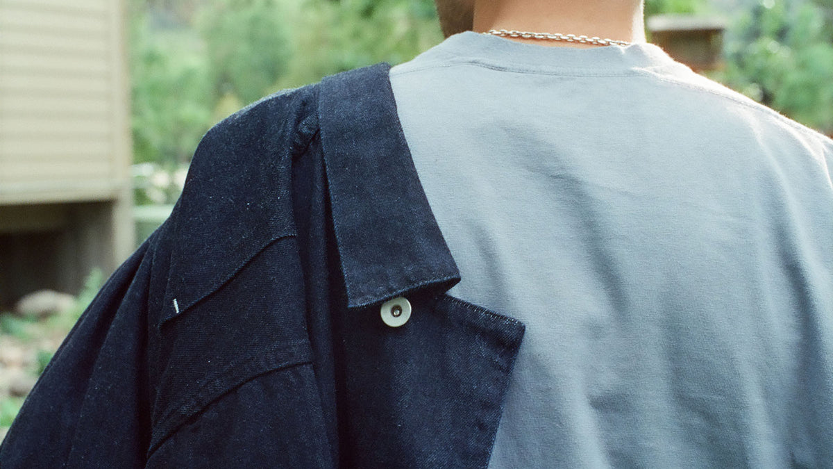 A person shown from behind wearing a light blue t-shirt and carrying a dark navy blue denim jacket over one shoulder in an outdoor setting.