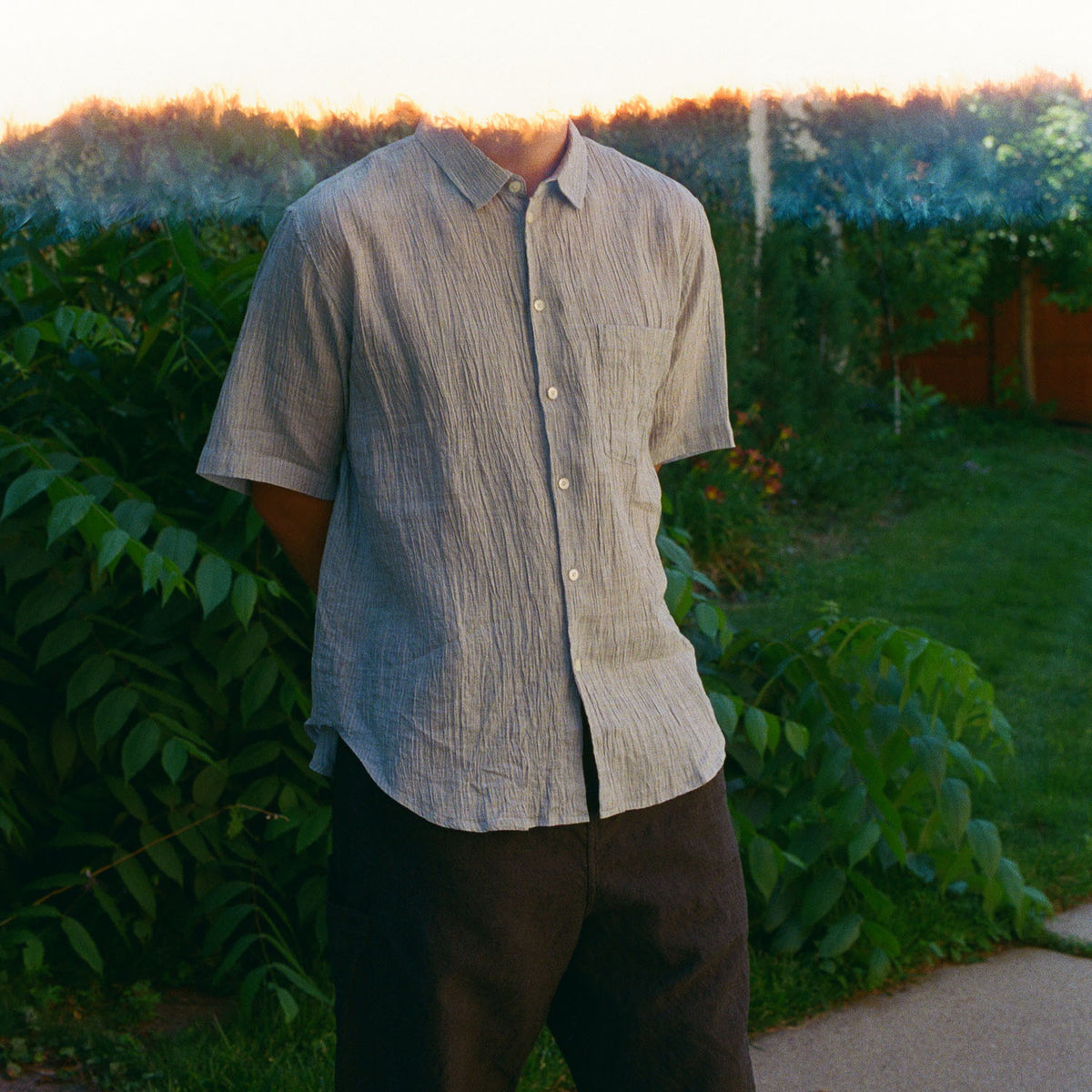 A person outdoors wearing a light gray short-sleeve button-up shirt with a subtle vertical textured weave and dark brown pants, standing in a garden setting with green foliage and warm sunset light.