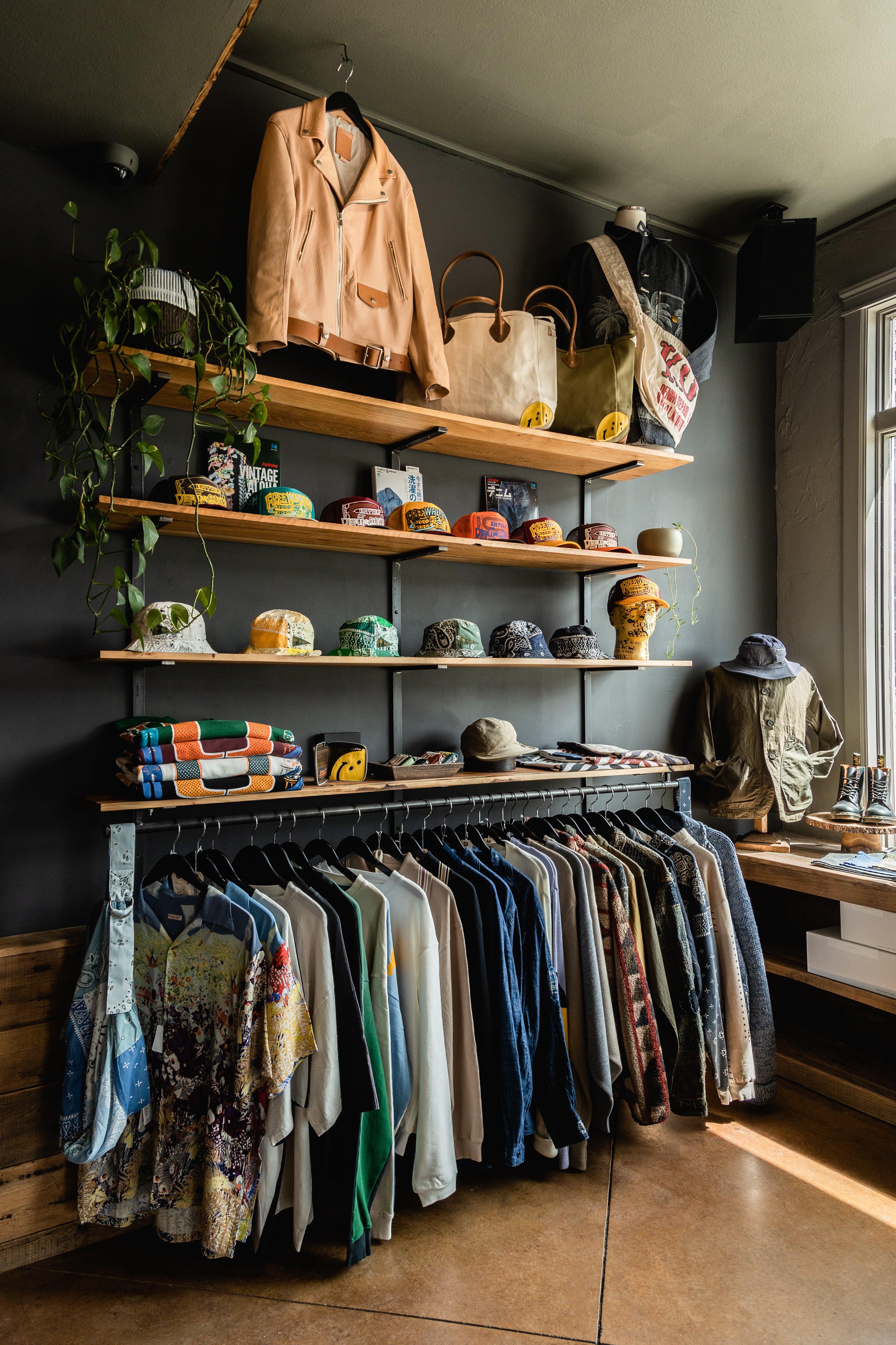 Interior photo of a Kapital display inside Canoe Club. There are wooden shelves lining the wall with Kapital Trucker hats and a green ivy plant. The lower rack ha hanging items from Kapital like button up shirts and jackets.