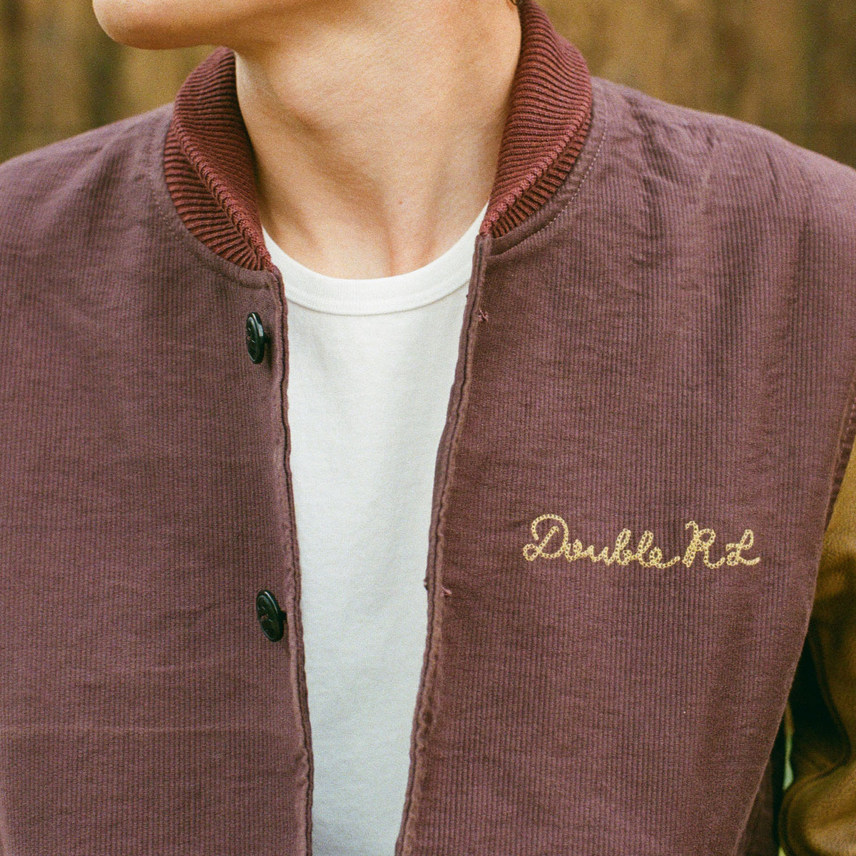 A close-up of a person wearing a corduroy varsity-style jacket in muted mauve-brown, layered over a white T-shirt. The jacket has a ribbed collar, dark buttons, and 'Double Rd' embroidered in gold on the chest.