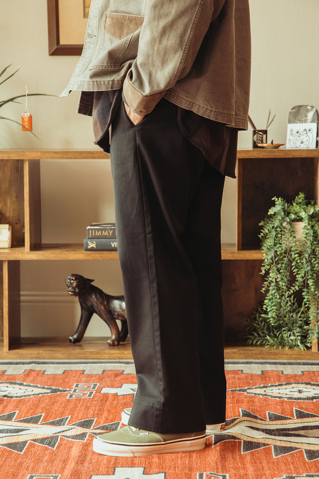 A person wearing loose black wide-leg trousers, a washed black jacket, and light green sneakers, standing on a patterned rug in front of a wooden shelf with plants and decor.
